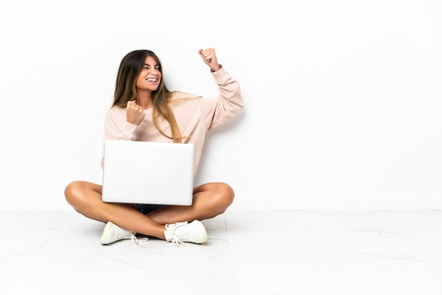 Jonge vrouw met een laptop zittend op de vloer geïsoleerd op een witte achtergrond een overwinning vieren