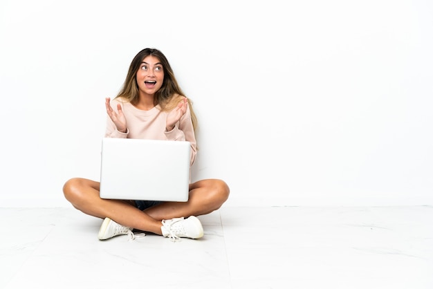 Jonge vrouw met een laptop zittend op de vloer geïsoleerd op een witte achtergrond met verrassingsgelaatsuitdrukking