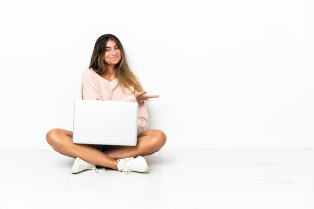 Jonge vrouw met een laptop zittend op de vloer geïsoleerd op een witte muur met een idee terwijl u glimlachend naar kijkt