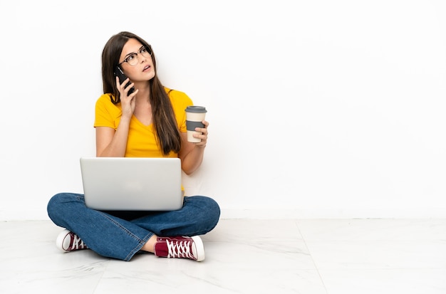 Jonge vrouw met een laptop zittend op de vloer met koffie om mee te nemen en een mobiel