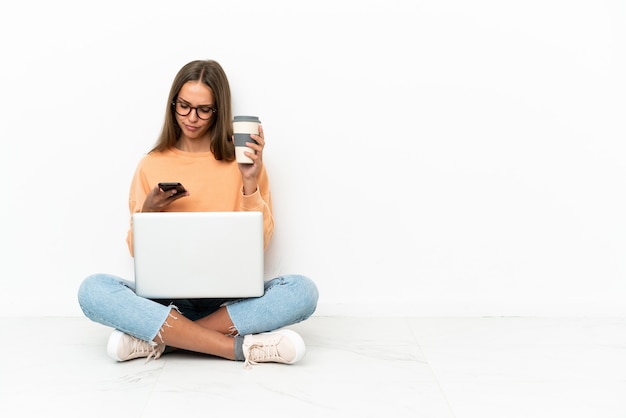 Jonge vrouw met een laptop zittend op de vloer met koffie om mee te nemen en een mobiel