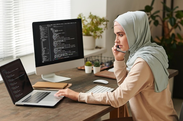 Jonge vrouw met een lichtblauwe hijab die aan tafel zit en belangrijke kwesties aan de telefoon bespreekt terwijl ze aan het werk is