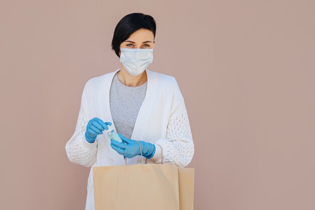 Jonge vrouw met een medisch wegwerpmasker en handschoenen met een papieren zak met aankopen tijdens de uitbraak van Covid 19. Levering dienstverleningsconcept.