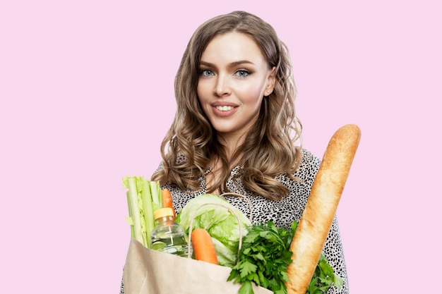 Jonge vrouw met een papieren zak met boodschappen. Mooie lachende blondine. Gezonde voeding, vitamines en veganisme. Geïsoleerd op een roze achtergrond.