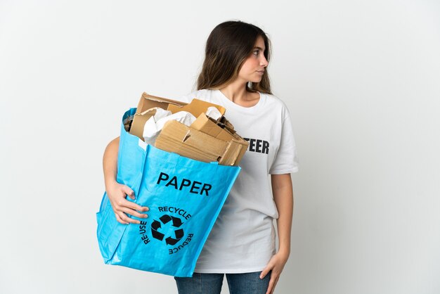 Jonge vrouw met een recycling zak vol papier om te recyclen geïsoleerd op een witte muur op zoek naar de zijkant