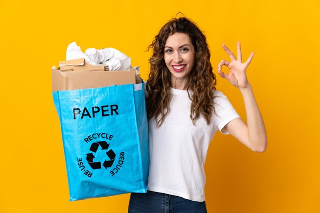 Jonge vrouw met een recycling zak vol papier om te recyclen geïsoleerd op gele muur met ok teken met vingers