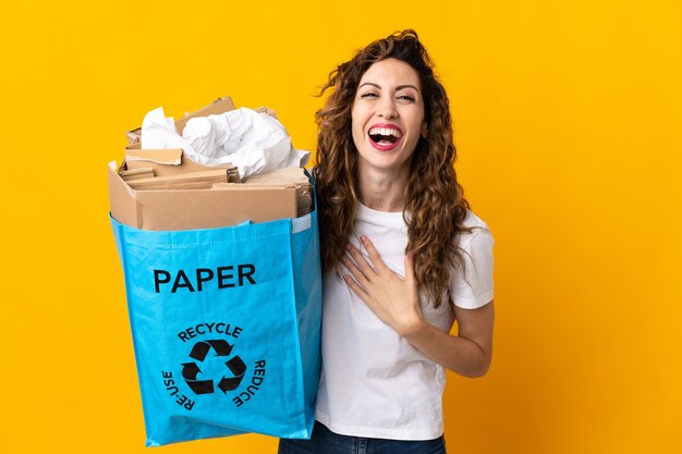 Jonge vrouw met een recyclingzak vol papier om te recyclen geïsoleerd op geel lachend veel