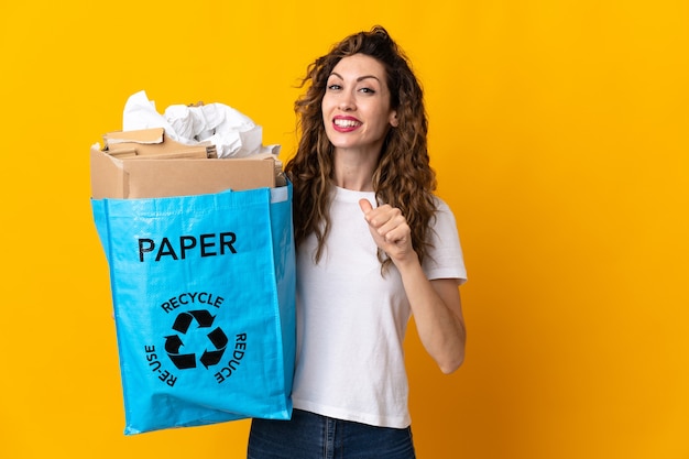 Jonge vrouw met een recyclingzak vol papier om te recyclen geïsoleerd op gele muur trots en zelfvoldaan