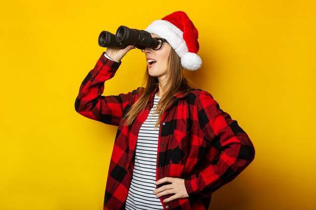 Jonge vrouw met een verbaasd gezicht in een kerstmuts en een rood shirt in een kooi kijkt door een verrekijker