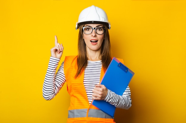 Jonge vrouw met een verbaasd gezicht in een vest en bouwvakker houdt een klembord op geel