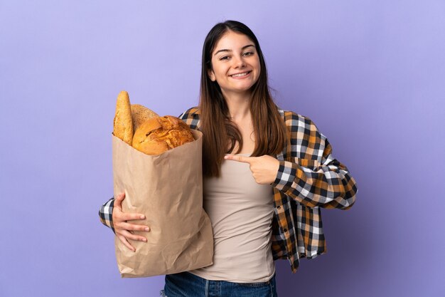 Jonge vrouw met een zak vol brood geïsoleerd op paars en wijst erop