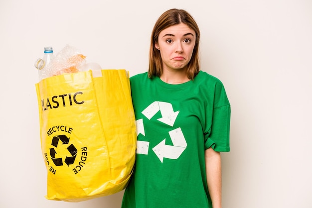 Jonge vrouw met een zak vol plastic flessen om te recyclen geïsoleerd op een witte achtergrond haalt haar schouders op en opent verwarde ogen