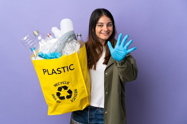 Jonge vrouw met een zak vol plastic flessen om te recyclen geïsoleerd op paars vijf tellen met vingers