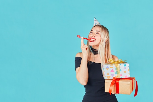 Jonge vrouw met geschenkdoos en blazende feesthoorn of lawaaimaker tijdens het vieren van verjaardag op een blauwe muur