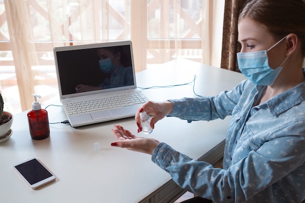 Jonge vrouw met gezichtsmasker die gadgets op haar werkplek desinfecteert