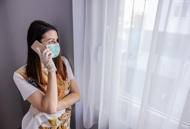 Jonge vrouw met gezichtsmasker en handschoenen die op mobiele telefoon spreken. F.