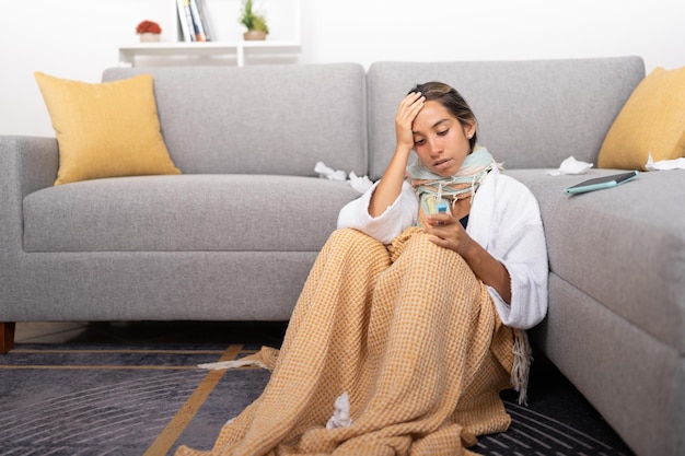 Jonge vrouw met griep en koorts zittend op de vloer van de woonkamer kijkend naar de thermometer