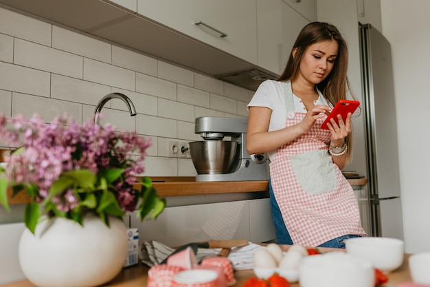 Jonge vrouw met groenten die lacht tijdens het gebruik van mobiele telefoon in de keuken