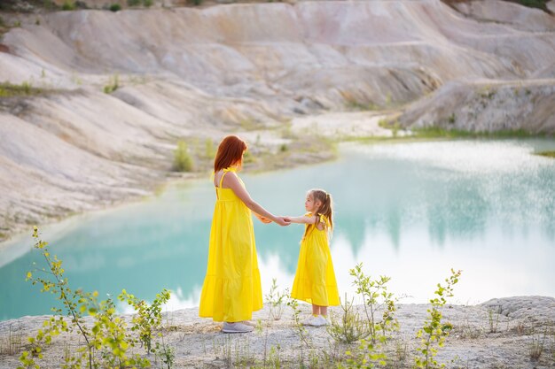 Jonge vrouw met haar dochter in een gele jurk in de buurt van het meer met azuurblauw water en groene bomen. gelukkig familie relatie concept