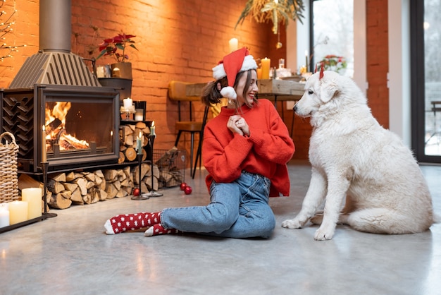 Jonge vrouw met haar schattige witte hond tijdens een gelukkig nieuwjaarsvakantie die thuis bij een open haard zit