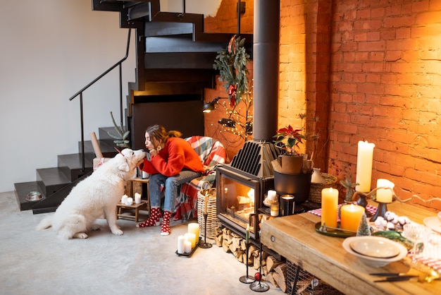 Jonge vrouw met haar schattige witte hond tijdens een gelukkig nieuwjaarsvakantie die thuis bij een open haard zit