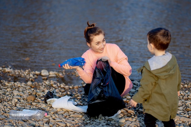 Jonge vrouw met haar zoon verzamelt plastic afval in een vuilniszak aan de rivier Lege gebruikte vuile plastic flessen Milieuvervuiling kust