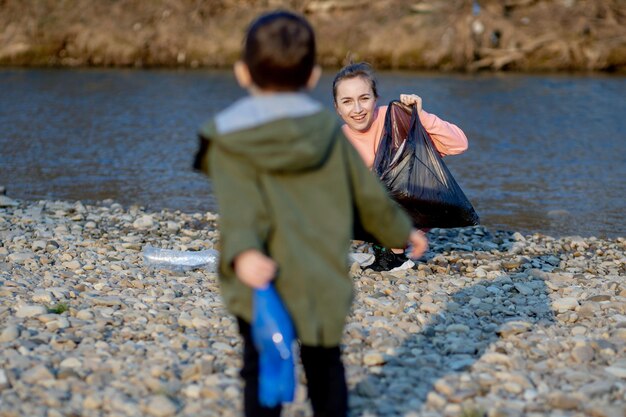 Jonge vrouw met haar zoon verzamelt plastic afval in een vuilniszak aan de rivier Lege gebruikte vuile plastic flessen Milieuvervuiling kust