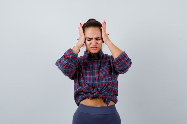 Jonge vrouw met handen op het hoofd in geruit hemd, broek en vermoeid, vooraanzicht.