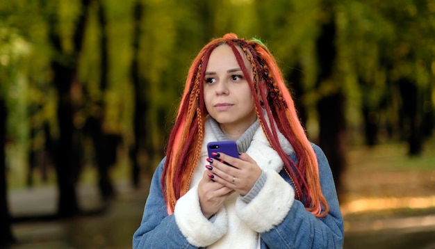 Jonge vrouw met heldere dreadlocks die mobiele telefoon doorbladert die zich in bospark bevindt Levendige vrouw met veelkleurig kapsel die smartphone op wandeling gebruikt