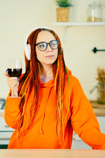 Jonge vrouw met heldere dreadlocks in glazen draadloze koptelefoon met glas rode wijn in de keuken