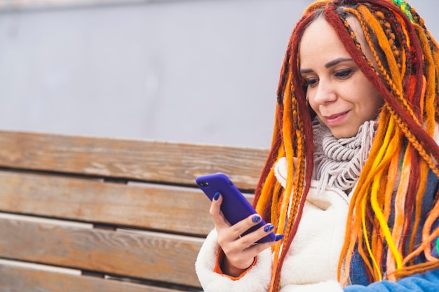 Jonge vrouw met heldere dreadlocks met mobiele telefoon op wandeling in het park Portret van een positieve vrouw in warme kleren die op smartphone bladert