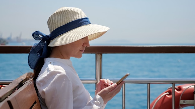 Jonge vrouw met hoed staat op het scheepsdek en surft op internet op smartphone tegen wazige zeiljachten in blauwe zee close-up