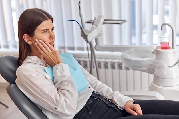 Jonge vrouw met kiespijn, zittend in een tandartsstoel in de kliniek. Vrouwelijke patiënt met vreselijke kiespijn die tandarts bezoekt.