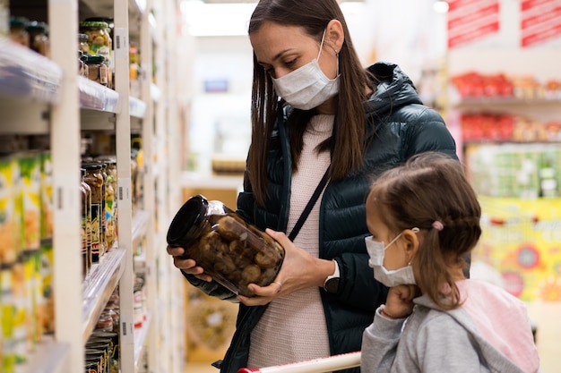 Foto jonge vrouw met kindmeisje in medische maskers koopt ingeblikt voedsel in kruidenierswinkelafdeling a