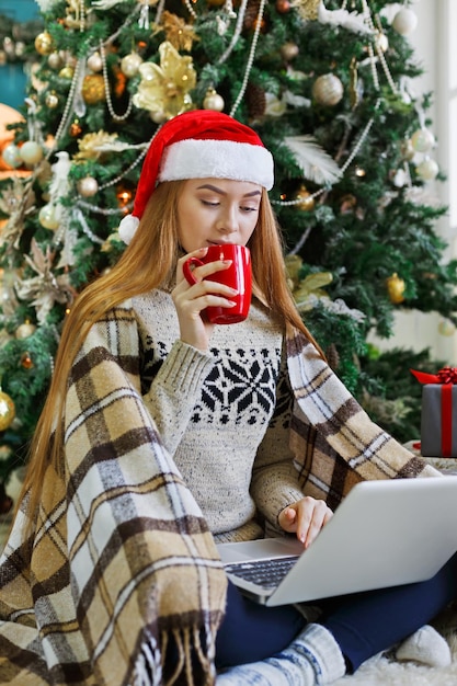 Jonge vrouw met koffie online winkelen op laptop in kerst interieur. Meisje zit onder versierde dennenboom tussen veel ingepakte cadeautjes. Voorbereiden op Kerstmis, op winterverkoop, kopieer ruimte