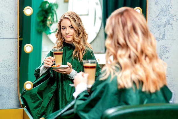 Jonge vrouw met kopje koffie latte spiegel in de schoonheidssalon kijken.