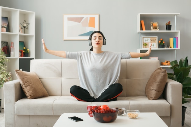 Jonge vrouw met koptelefoon die yoga doet, zittend op de bank achter de salontafel in de woonkamer