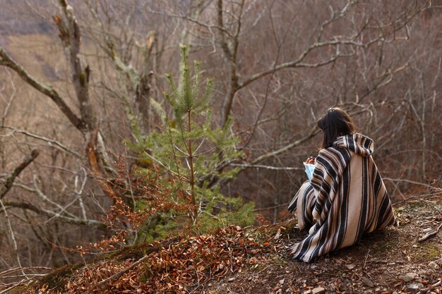 Jonge vrouw met kort donkerbruin haar in poncho zetel neer in de herfstbossen, bos.