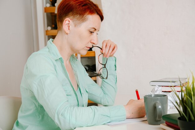 Jonge vrouw met kort haar aan het bureau schrijft bedachtzaam in rood potlood Verfrommeld papier op het bureau