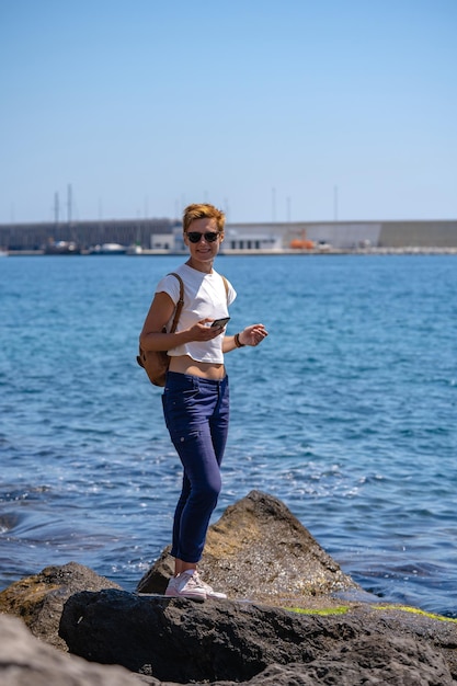 Jonge vrouw met kort haar en rugzak aan de kust van de Middellandse Zee met vuurtoren en boten op de achtergrond