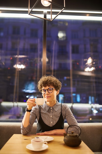 Jonge vrouw met kort krullend haar zitten bij tafel in café, ontspannen en met thee of koffie