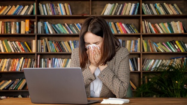 Jonge vrouw met kort los haar niest en neemt wit papieren servet zittend op grijze laptop tegen gekleurde boekenplanken in kantoor