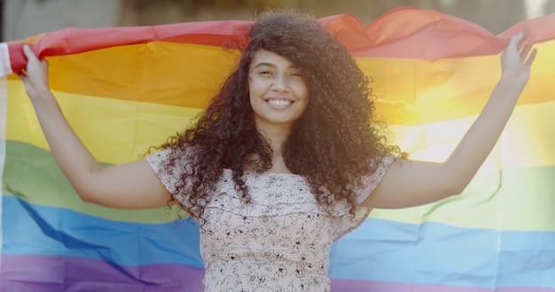 jonge vrouw met krullend haar bedekt met lgbt-trotsvlag die vuist omhoog houdt die lgbt-vlag bedekt