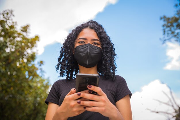 Foto jonge vrouw met krullend haar gebruikt haar telefoon, ze heeft een zwart masker op haar gezicht