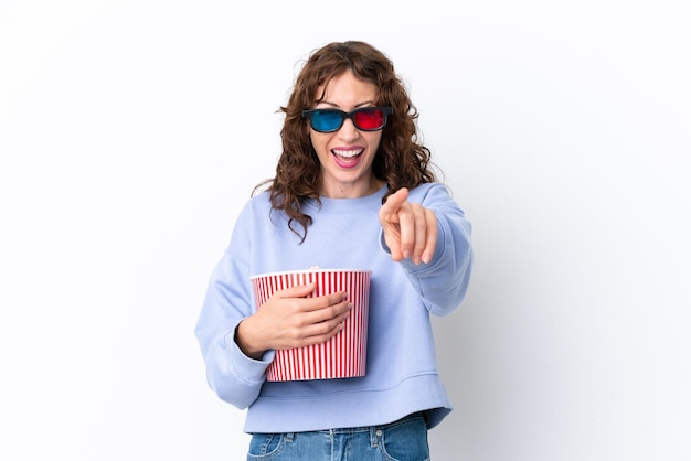 Jonge vrouw met krullend haar geïsoleerd op een witte achtergrond met 3d-bril en met een grote emmer popcorns terwijl ze naar voren wijst