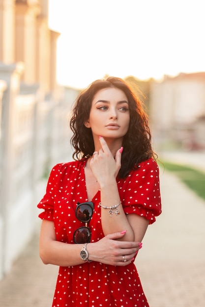 Jonge vrouw met krullend haar met modieuze sieradenarmband en horloge in rode jurk met stippen loopt in de stad bij zonsondergang