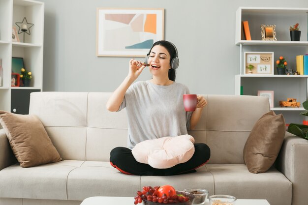 Jonge vrouw met kussen met koptelefoon eet koekjes met een kopje thee op de bank achter de salontafel in de woonkamer