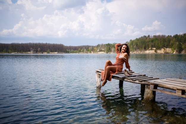Jonge vrouw met lang golvend krullend haar in een lange guipurejurk op blote voeten in de zomer in een bos aan een meer bij zonsondergang staande op een pantone op een houten pierbrug. Zomer zonnige dag