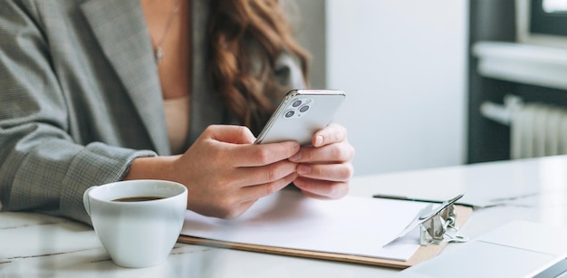 Jonge vrouw met lang haar in stijlvol pak met behulp van mobiele telefoon in handen werken in de heldere moderne kantoorvrouw bericht verzenden via smartphone