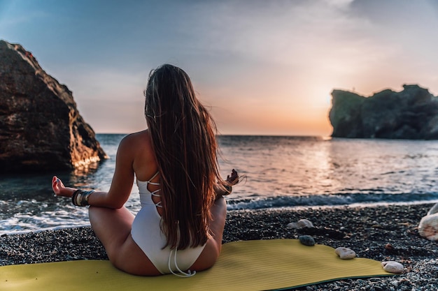 Jonge vrouw met lang haar in wit zwempak en boho-stijl armbanden die buiten op yogamat aan zee oefenen op een zonsondergang Vrouwen yoga fitness routine gezonde levensstijl harmonie en meditatie